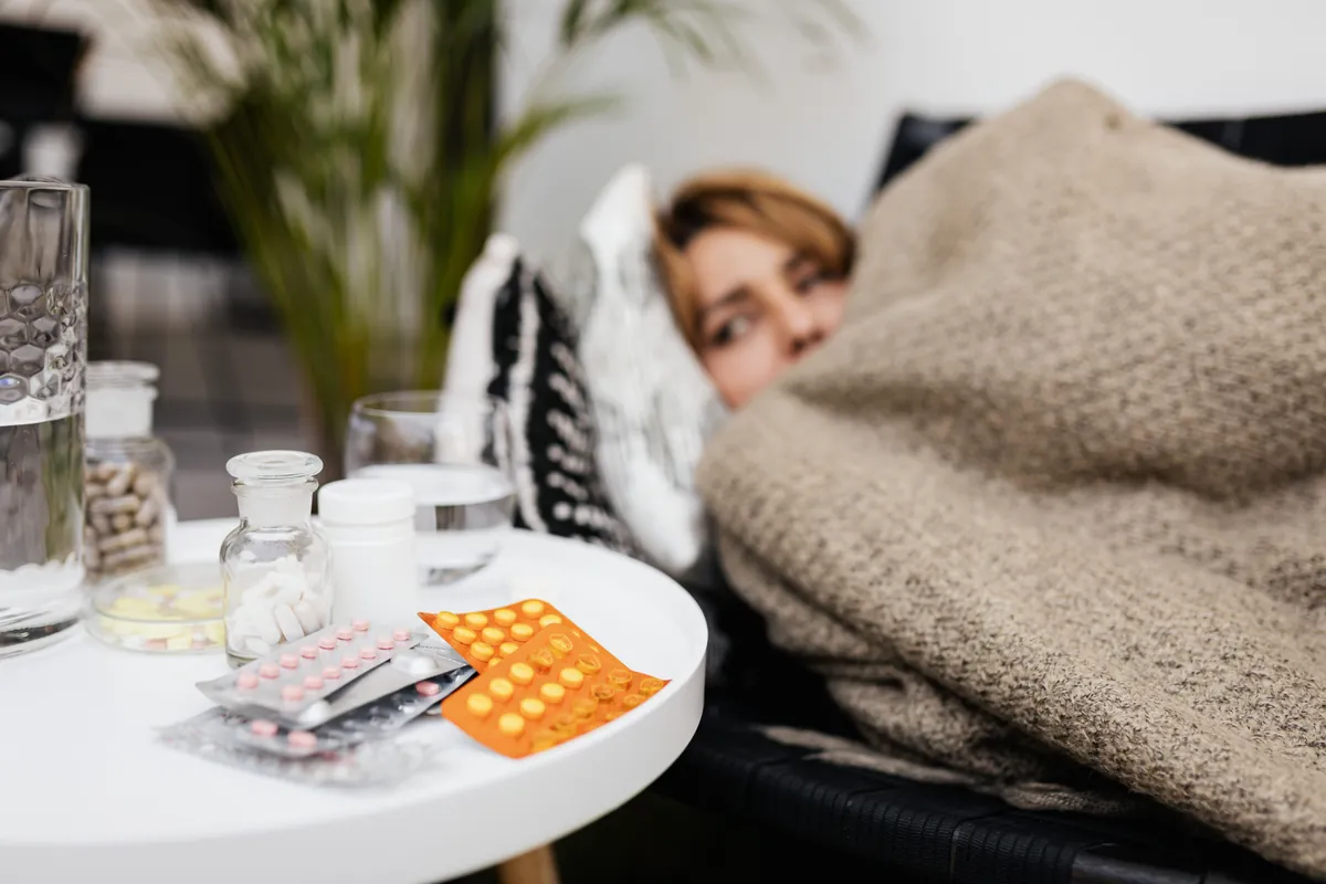 Una mujer acostada en el sillón, tapada con una cobija, junto a una mesita llena de pastillas y frascos y un vaso de agua.