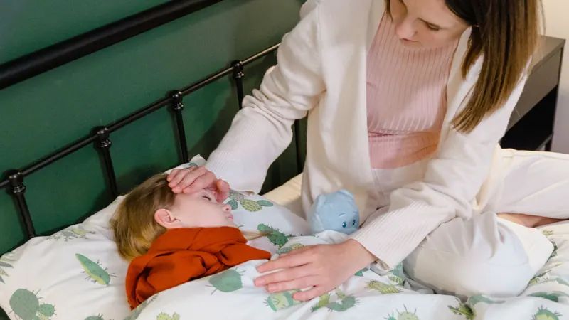 Una madre sentada en la cama con la mano en la frente de su hijo, que está acostado y arropado con cara de malestar.