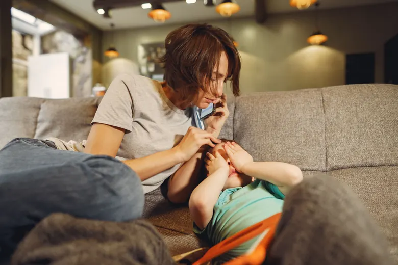 Una madre habla por teléfono sentada en el sillón de su sala, con la otra mano sobre la cabeza de su hijo, que está acostado junto a ella tapándose los ojos.